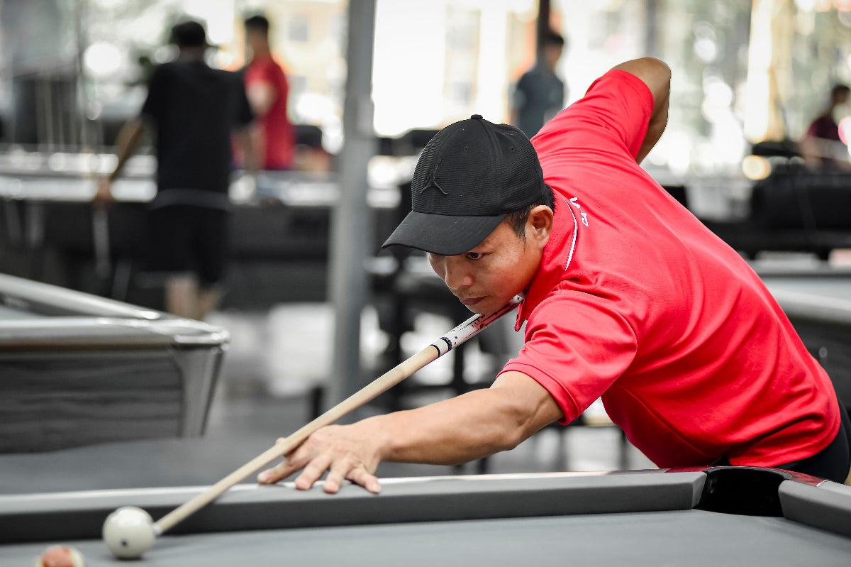 A man in a red polo shirt and black cap leans over a pool table, holding a pool cue with a white ball nearby, in a real indoor environment with blurred people in the background.