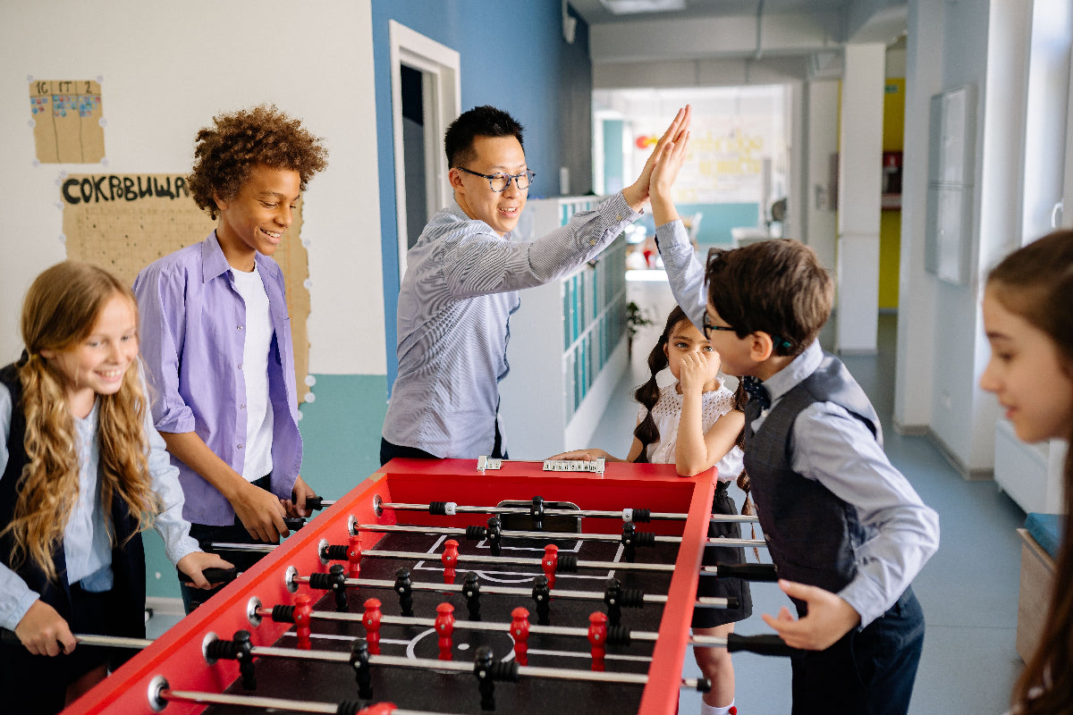 A group of children and an adult playing with a red foosball table in a bright indoor room. The scene is highly active with multiple people interacting around the table.