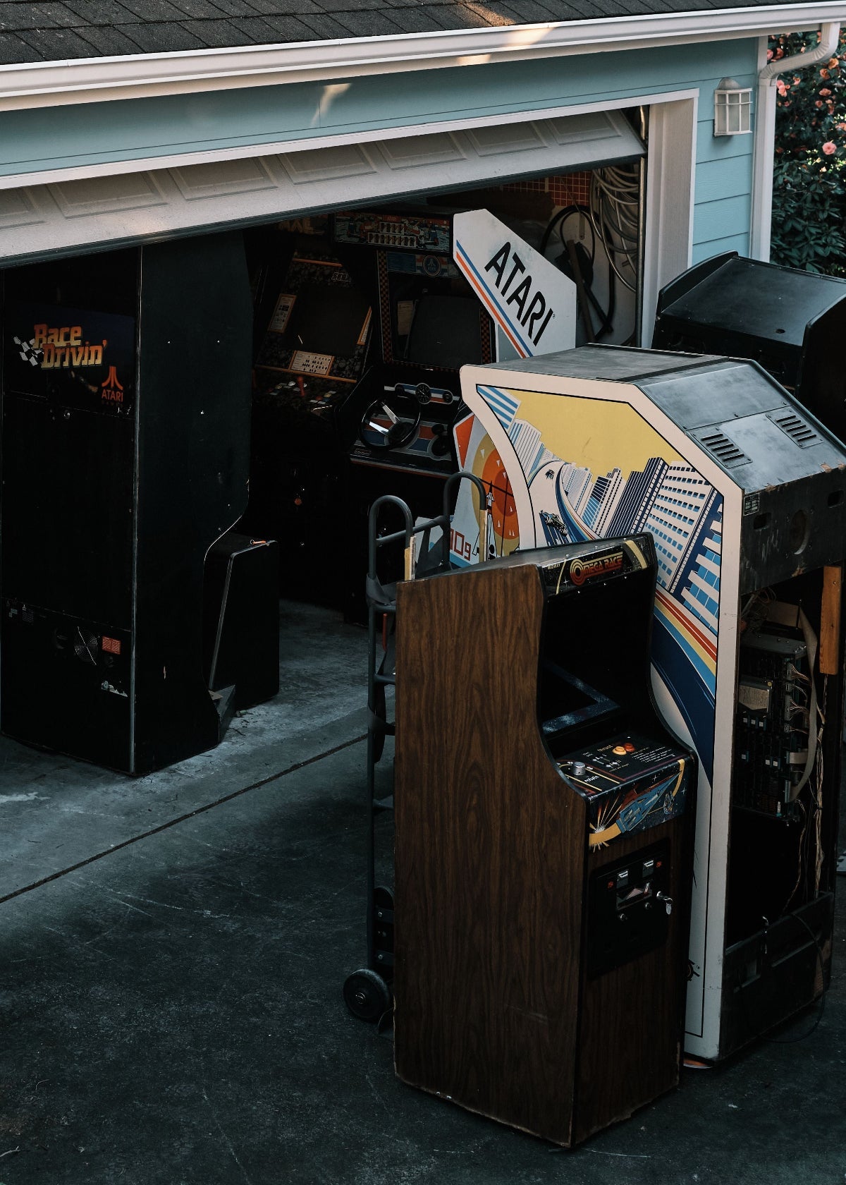 A vintage Atari arcade machine with a dark wood cabinet stands in front of an open garage door, next to other retro arcade machines.