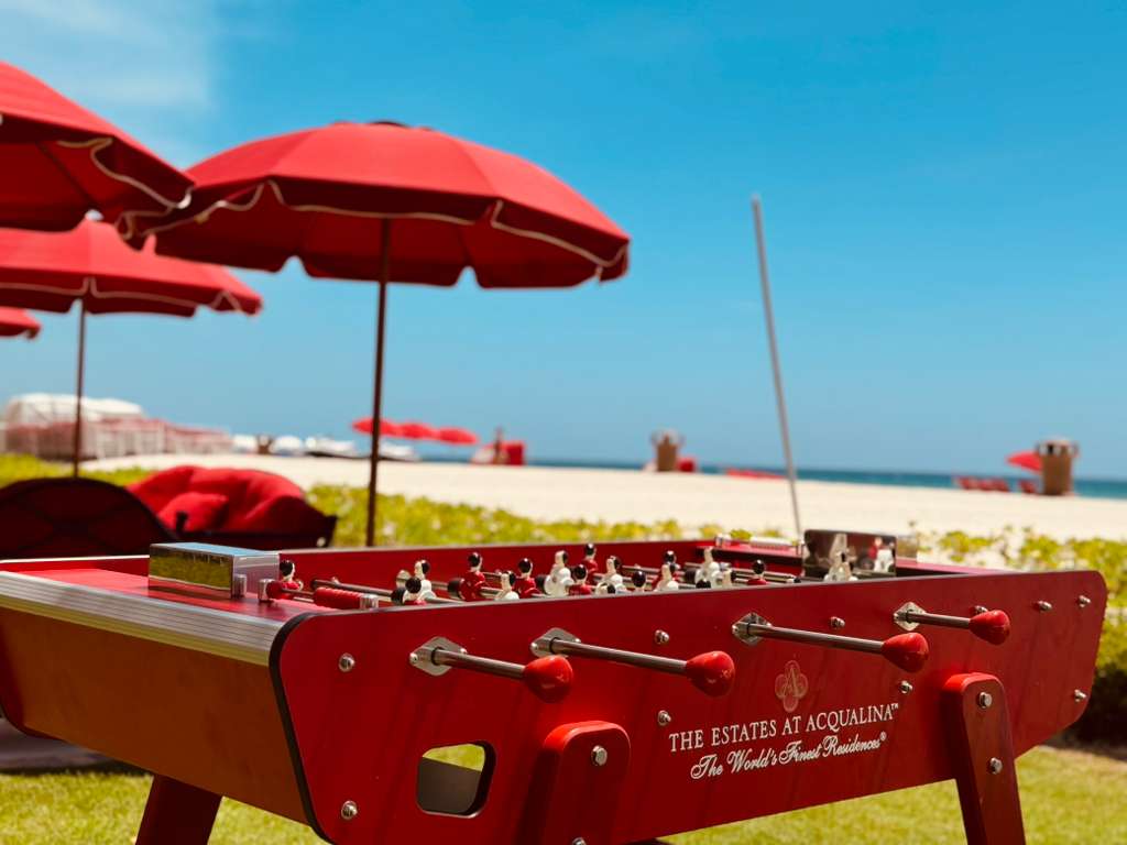 A red foosball table in the foreground with beach umbrellas, sand, and ocean in the background, branded 'THE ESTATES AT ACQUALINA'.