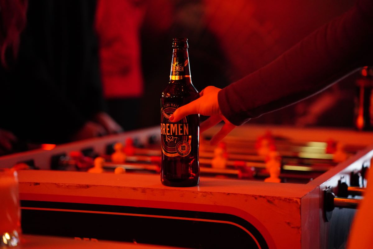 A hand holding a bottle of Bremen Kölsch beer under dramatic red ambient lighting, positioned over a foosball table in a dimly lit setting.