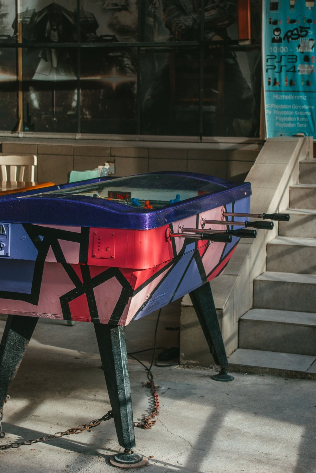 A colorful foosball table with a geometric graffiti-style pattern in pink, blue, and red, positioned next to concrete stairs in an indoor setting.