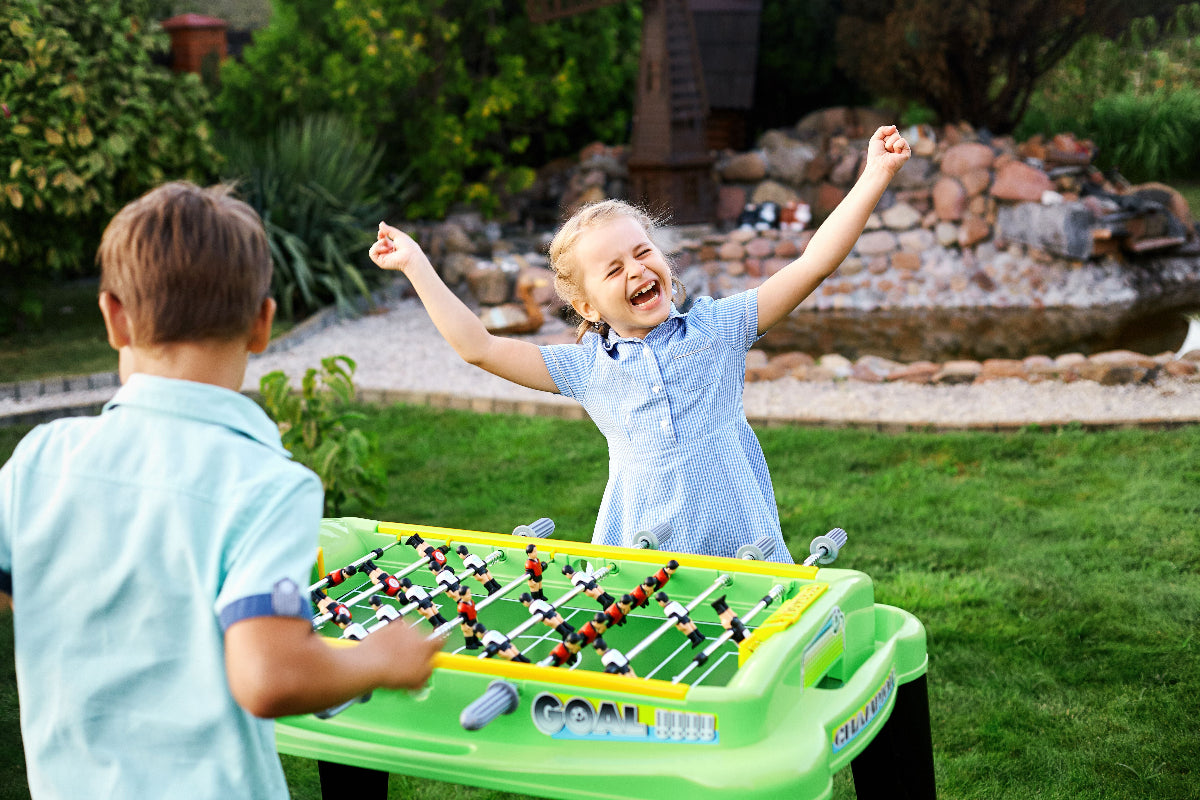 Two children playing with a bright green foosball table outdoors on a grassy lawn, with a garden and stone feature in the background.