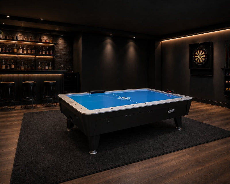 A air hockey table with a bright blue surface sits in a dimly lit home bar or recreation room with wood floors, a bar counter with stools, and a dartboard on the wall.