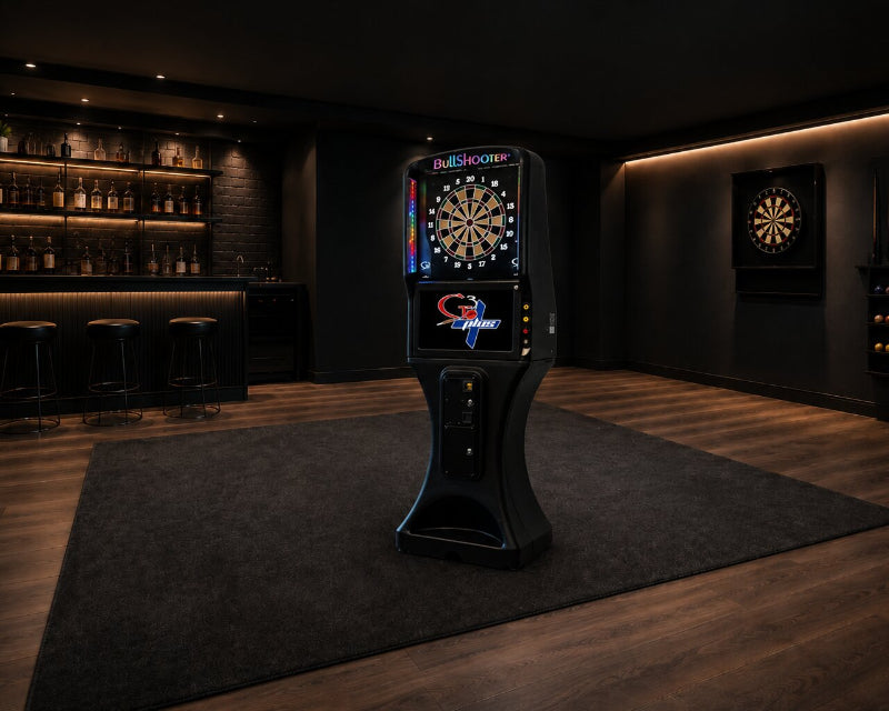A blackArachnid dartboard machine stands on a dark gray rug in a dimly lit home bar or gaming room with wood floors, shelves of bottles, and another dartboard on the wall.