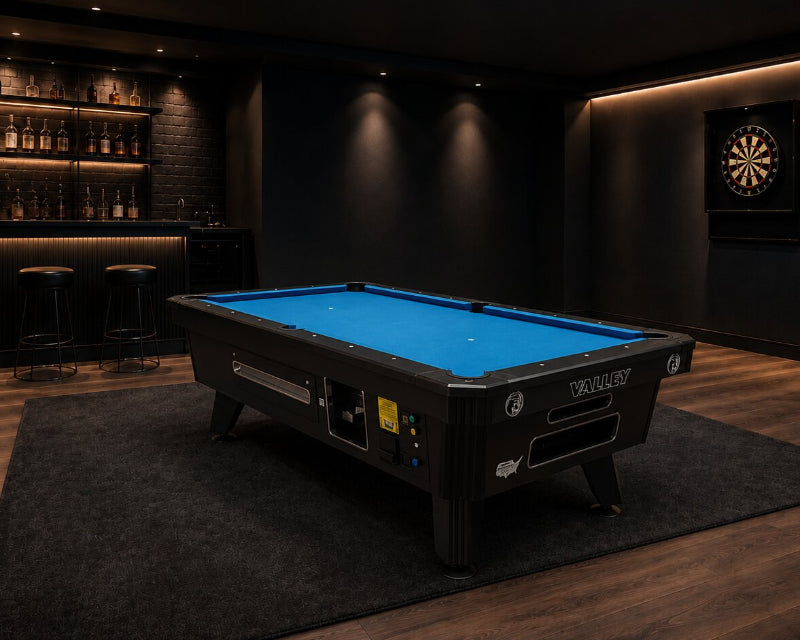 A black pool table with a bright blue felt surface is shown in a dimly lit home bar or recreation room with wood floors, shelves of bottles, a dartboard, and a rug underneath the table.